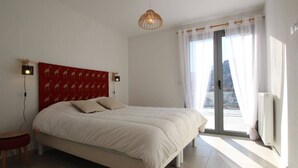 A bedroom with a bed, a red headboard, two bedside lamps, a small stool, and a view of the outside through a large window.