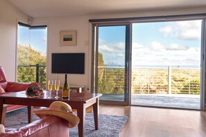 Un soggiorno moderno con un tavolo in legno, un divano e un'ampia porta scorrevole in vetro che dà su un balcone con vista su una recinzione e alberi.