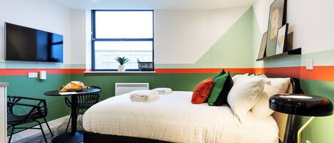 A hotel room with a bed, a TV, a window, a small table with chairs, and a radiator.