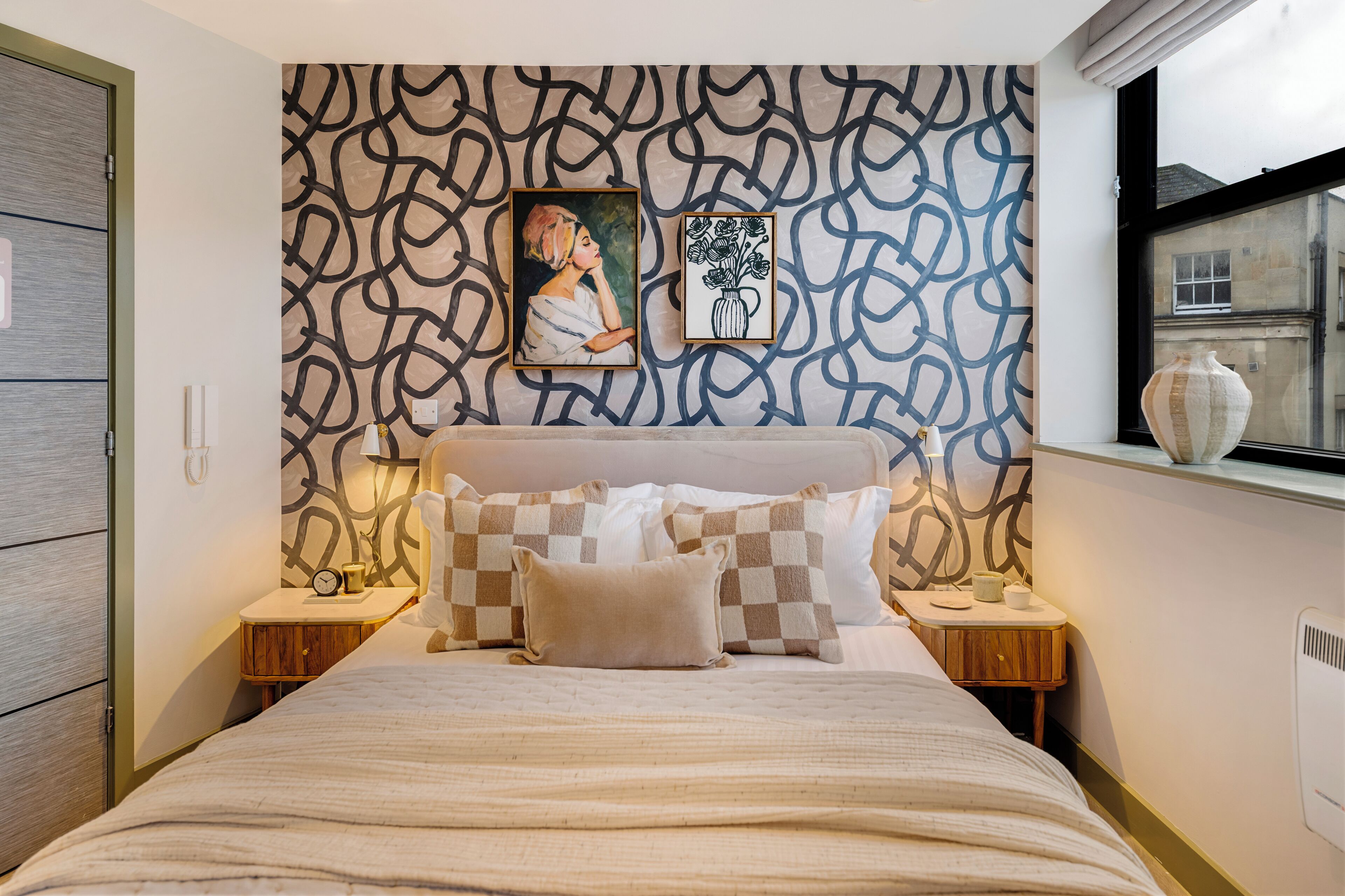 Une chambre avec un papier peint à motifs, un lit agrémenté d’oreillers à carreaux, une table de chevet avec une lampe, et une œuvre d’art encadrée au-dessus du lit.
