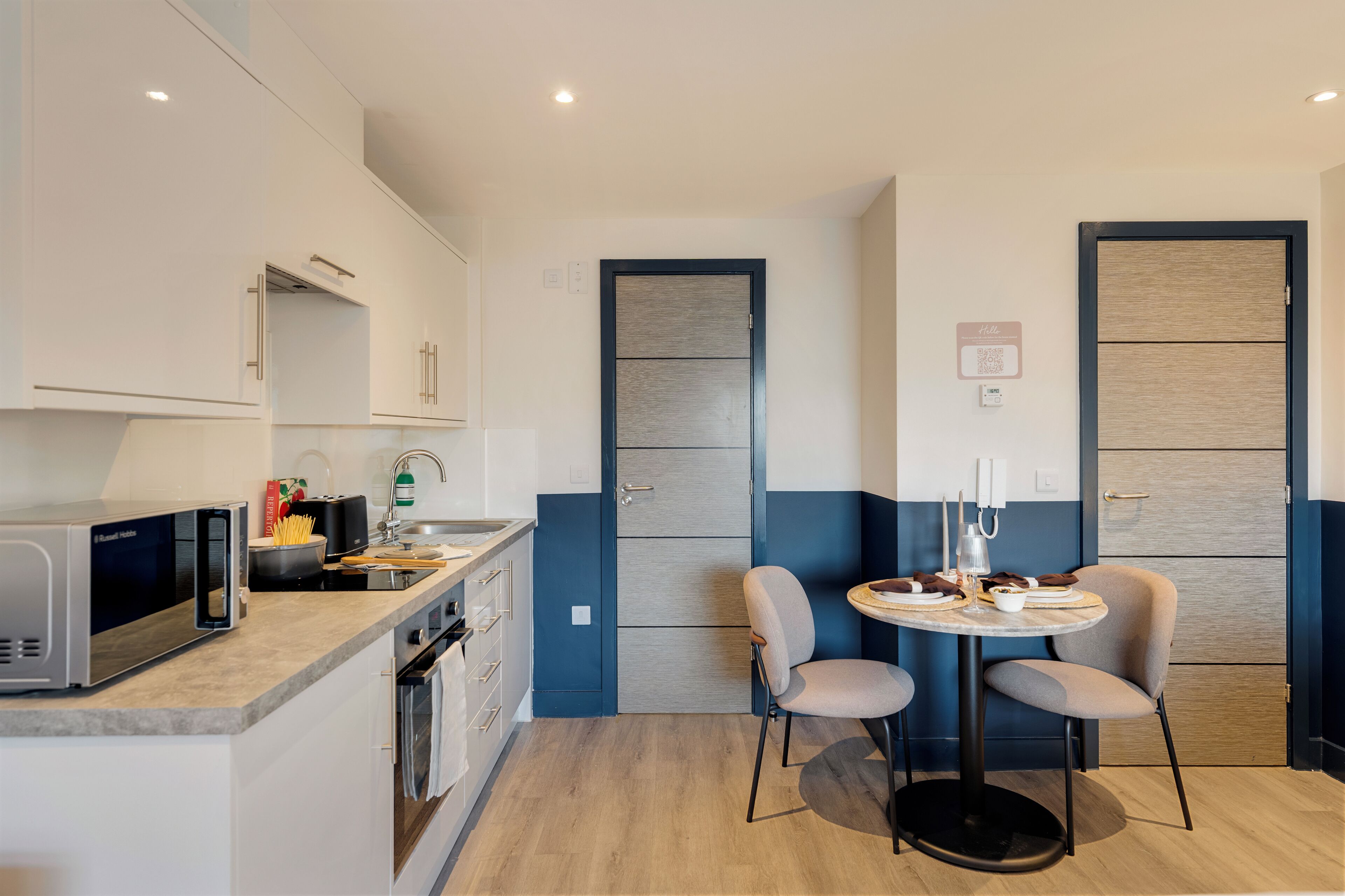 Una cocina compacta con microondas, lavamanos y armarios. Cuenta con un pequeño comedor con dos sillas y una mesa.