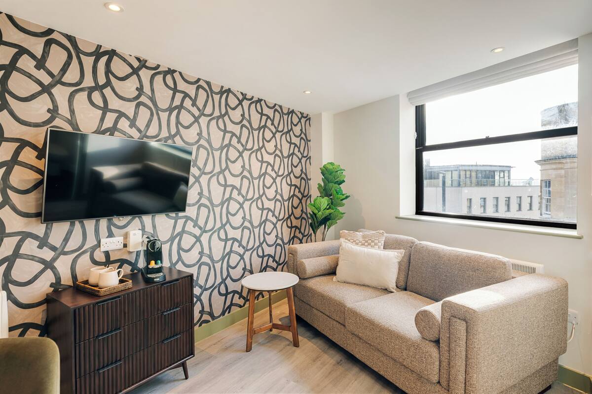 Ein modernes Wohnzimmer mit einer Couch, einem Couchtisch, einem an der Wand montierten Fernseher und einem Fenster mit Blick auf die Stadt.
