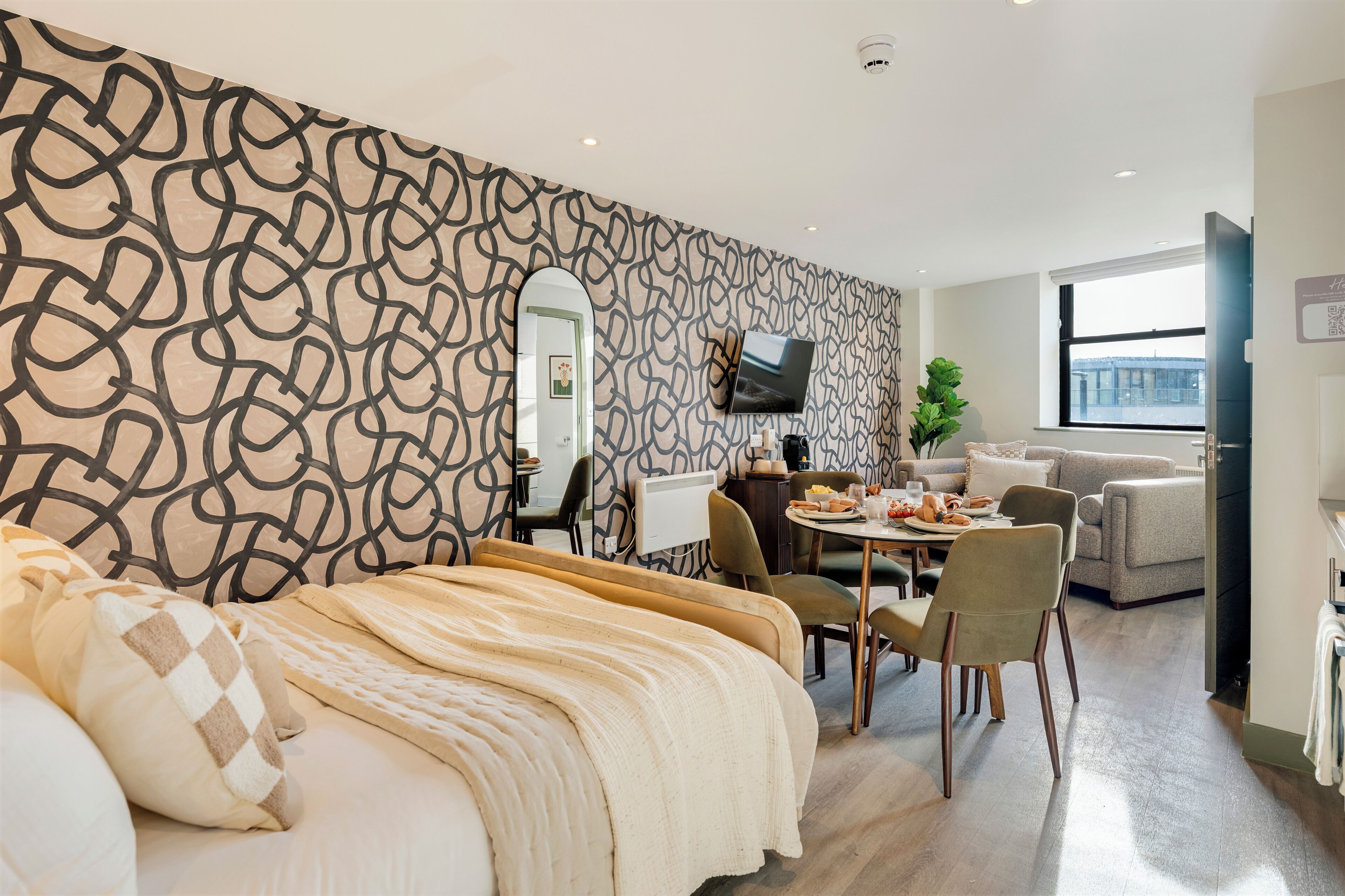 Una habitación de hotel moderna con una cama grande, un área de comedor con mesa y sillas, un televisor montado en la pared y una ventana con vista a la ciudad.