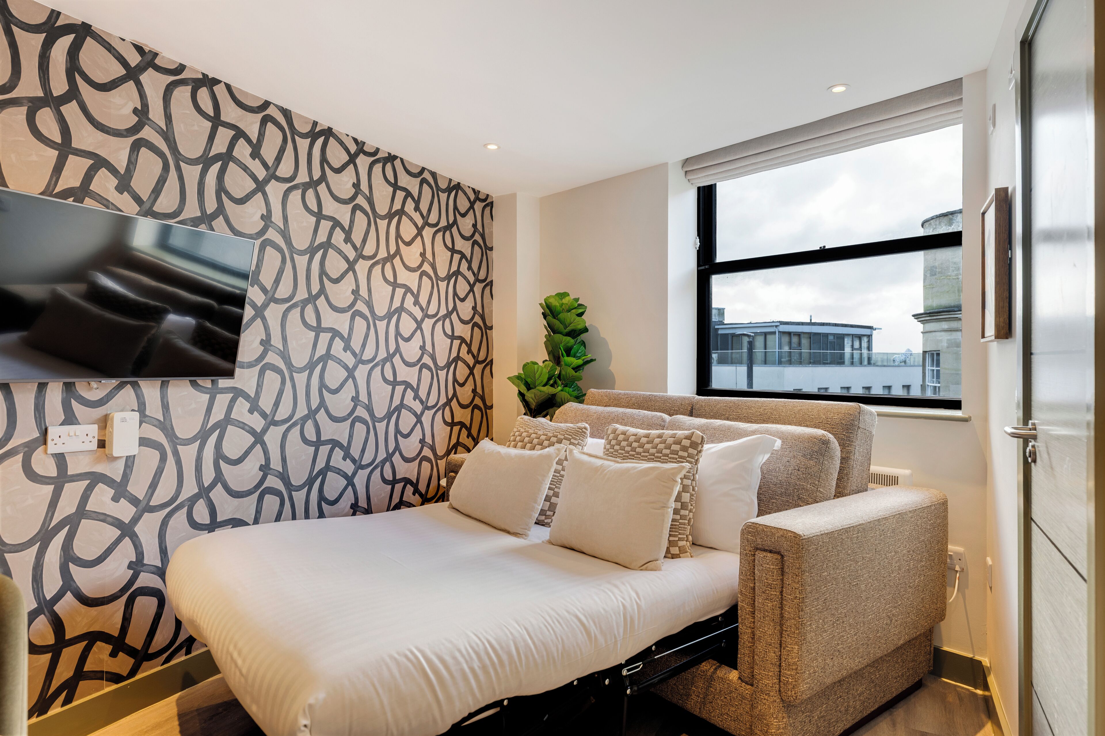 A modern bedroom with a bed, a TV mounted on the wall, a window with a view of buildings, and a patterned wall.