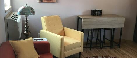Un salon chaleureux avec un canapé rouge, un fauteuil jaune, une petite table avec une télécommande, un téléviseur fixé au mur et un tapis à motifs.