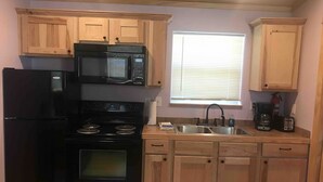 A compact kitchen with a black refrigerator, stove, and microwave, wooden cabinets, a window with blinds, and a sink with a faucet.