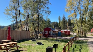 A picnic area with a wooden bench and a red umbrella, surrounded by trees and a red building. - Cabin #5 | 10 Min to Glacier Park Entrance, Minutes to Glacier Park! (Coram)