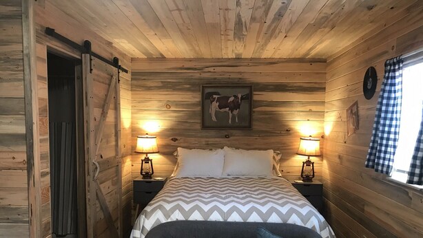 A wooden cabin bedroom with a bed, two bedside lamps, a clock, and a window with curtains. - Cabin #5 | 10 Min to Glacier Park Entrance, Minutes to Glacier Park! (Coram)