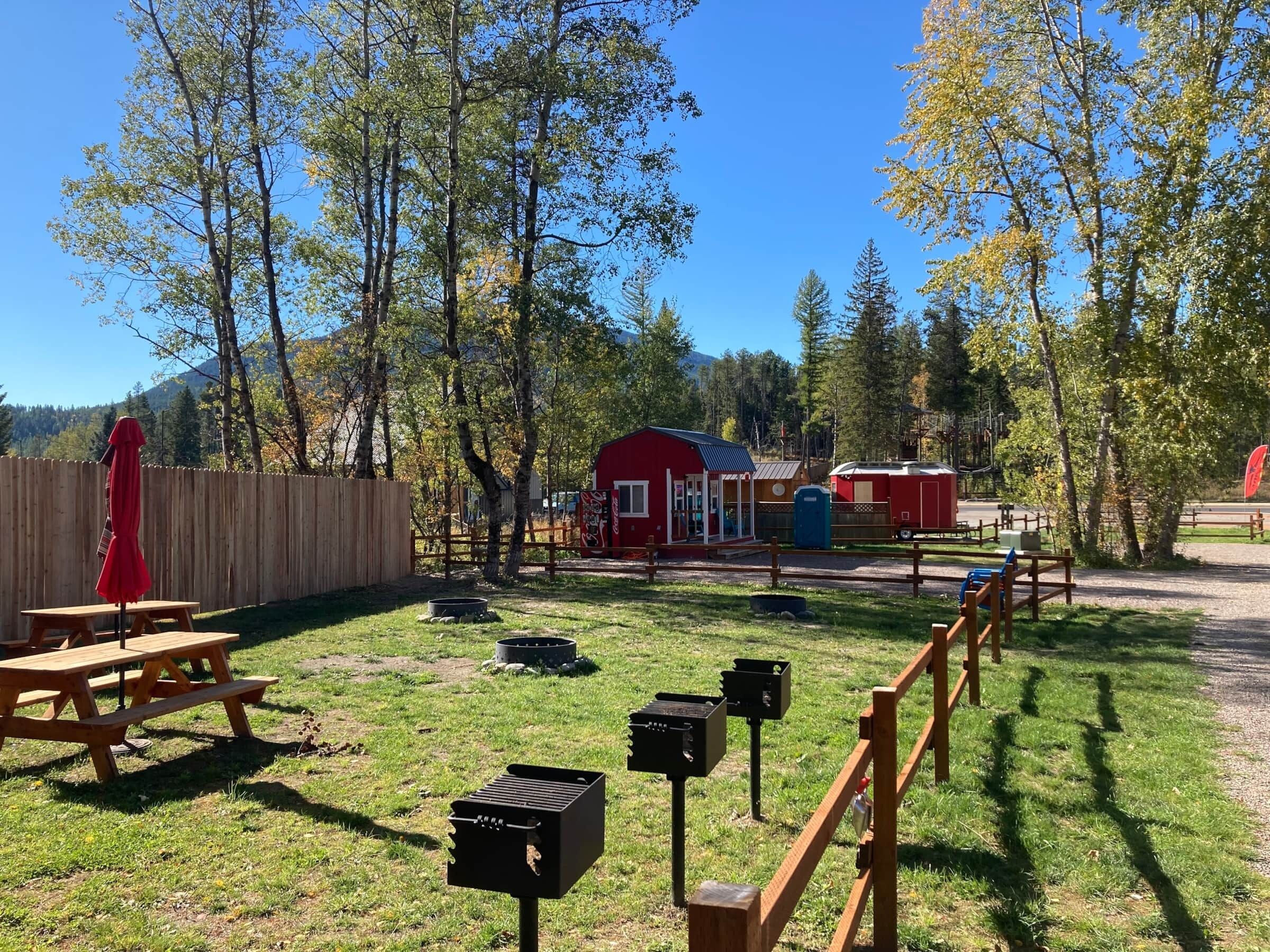 A picnic area with a wooden bench and a red umbrella, surrounded by trees and a red building.
