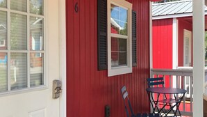 Fachada roja con una puerta blanca, una ventana con contraventanas y una pequeña mesa con sillas en una terraza de madera.