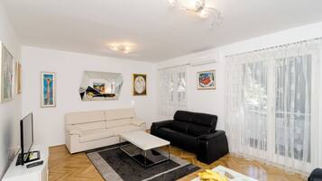 Una sala de estar con un sofá color beige, un sofá negro, una mesa de centro y un televisor sobre un mueble blanco. Se observan cuadros enmarcados en las paredes y una ventana con cortinas translúcidas.
