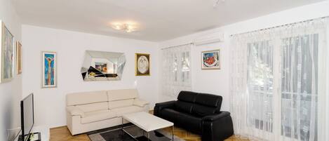 A living room with a beige sofa, a black sofa, a coffee table, and a TV on a white stand. There are framed pictures on the walls and a window with sheer curtains.