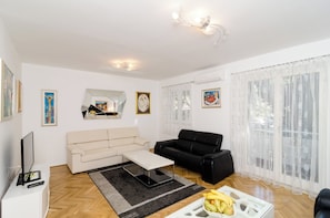 A living room with a beige sofa, a black sofa, a coffee table, and a TV on a white stand. There are framed pictures on the walls and a window with sheer curtains. - Apartment Iva (Dubrovnik)
