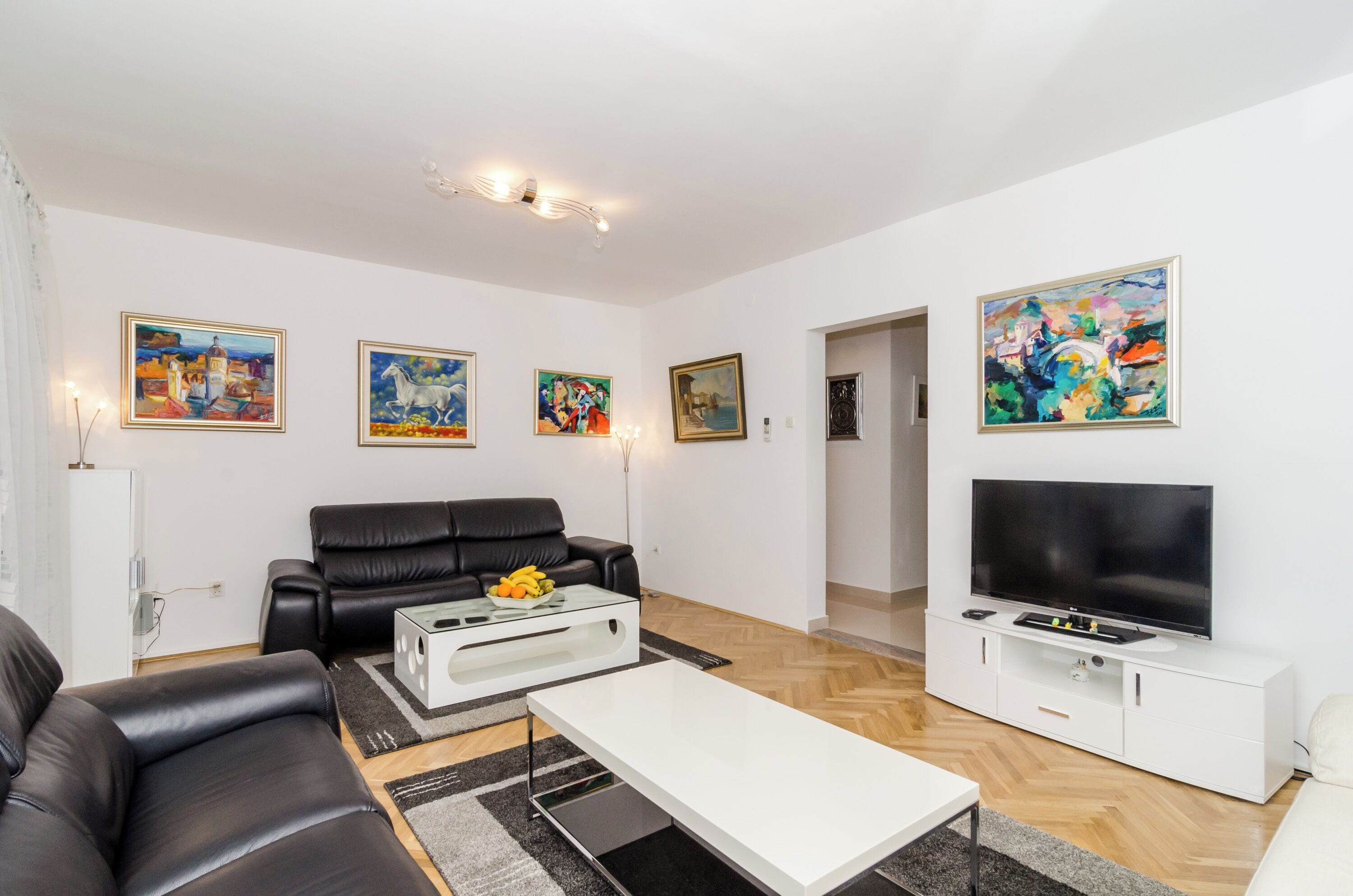 A modern living room with a black leather sofa, a white coffee table, a flat-screen TV on a white cabinet, and various paintings on the walls.