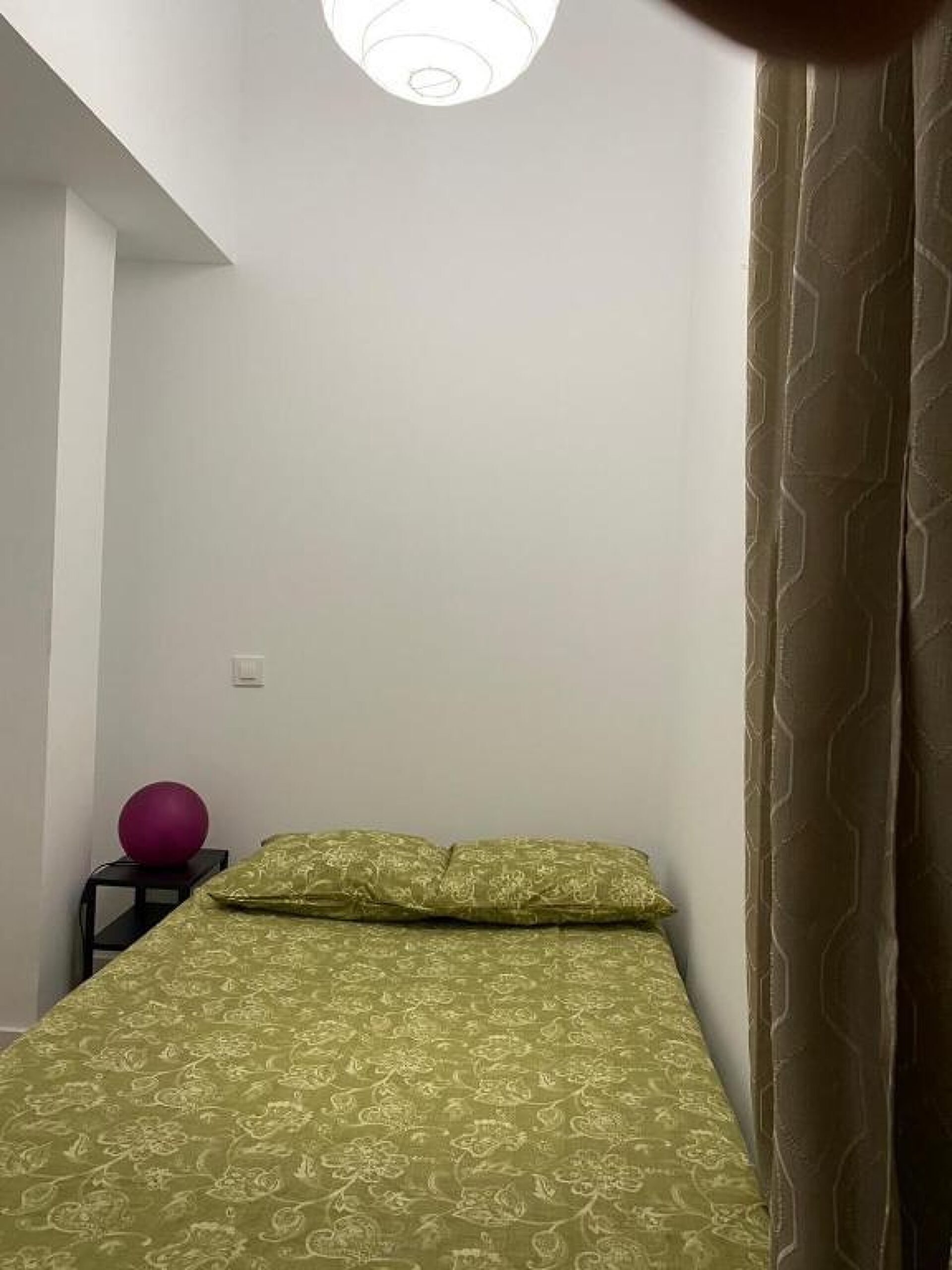Een kleine kamer met een enkel bed, een beddegoed met patroon, een stuiterbal en een gordijn met dessin.