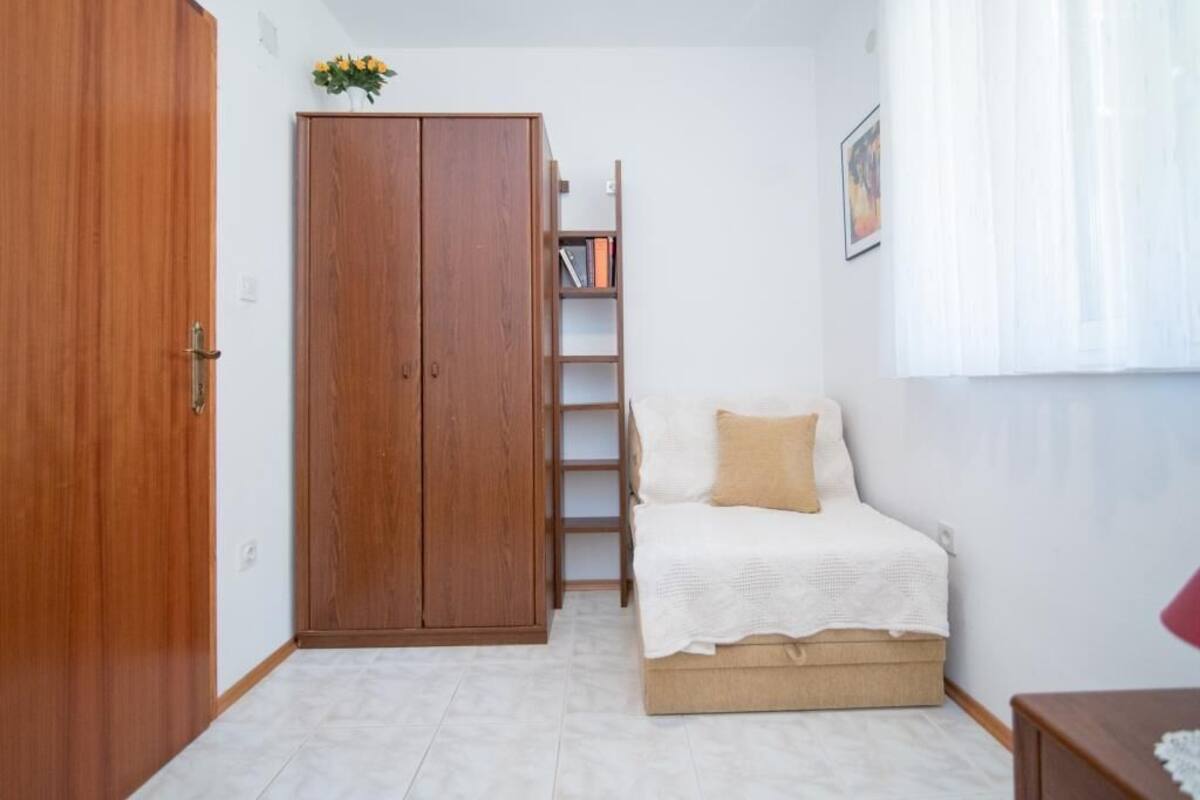 A bedroom with a wooden wardrobe, a bed with a white and beige duvet, a small bookshelf, and a window with sheer curtains.