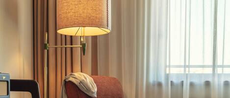 Une chambre d’hôtel avec un parquet en bois disposé en motif chevrons, un fauteuil rouge recouvert de tissu avec un coussin décoratif, une commode en bois dotée de nombreux tiroirs, une petite table et une lampe avec un abat-jour en osier.