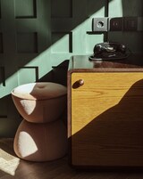 A wooden chest of drawers with a telephone on top, a cushioned stool, and a wall with geometric patterns.