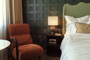 Una camera d'albergo con una testiera verde, una poltrona imbottita rossa, un comodino con una lampada e un letto rifatto con lenzuola bianche.