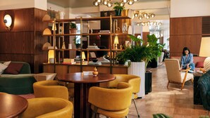A modern lobby with a wooden reception desk, yellow chairs, a round table, and a bookshelf filled with books and decorative items.