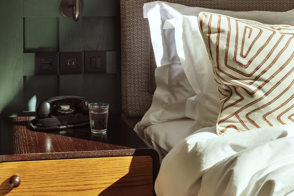 A neatly made bed with patterned bedding, a bedside lamp, a telephone, and a glass of water on a wooden nightstand.