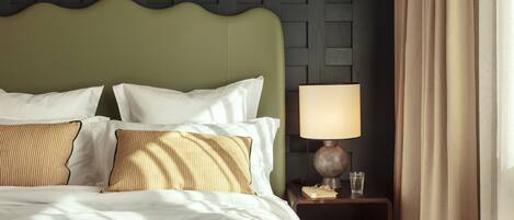 A neatly made bed with white and striped pillows, a green upholstered headboard, a bedside lamp, and a small bedside table with books and a glass.