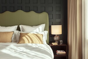 A neatly made bed with white and striped pillows, a green upholstered headboard, a bedside lamp, and a small bedside table with books and a glass.