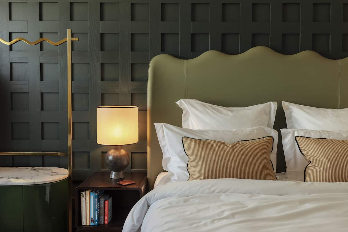 A neatly made bed with white linens and striped pillows, a bedside lamp, and a small bookshelf with books.