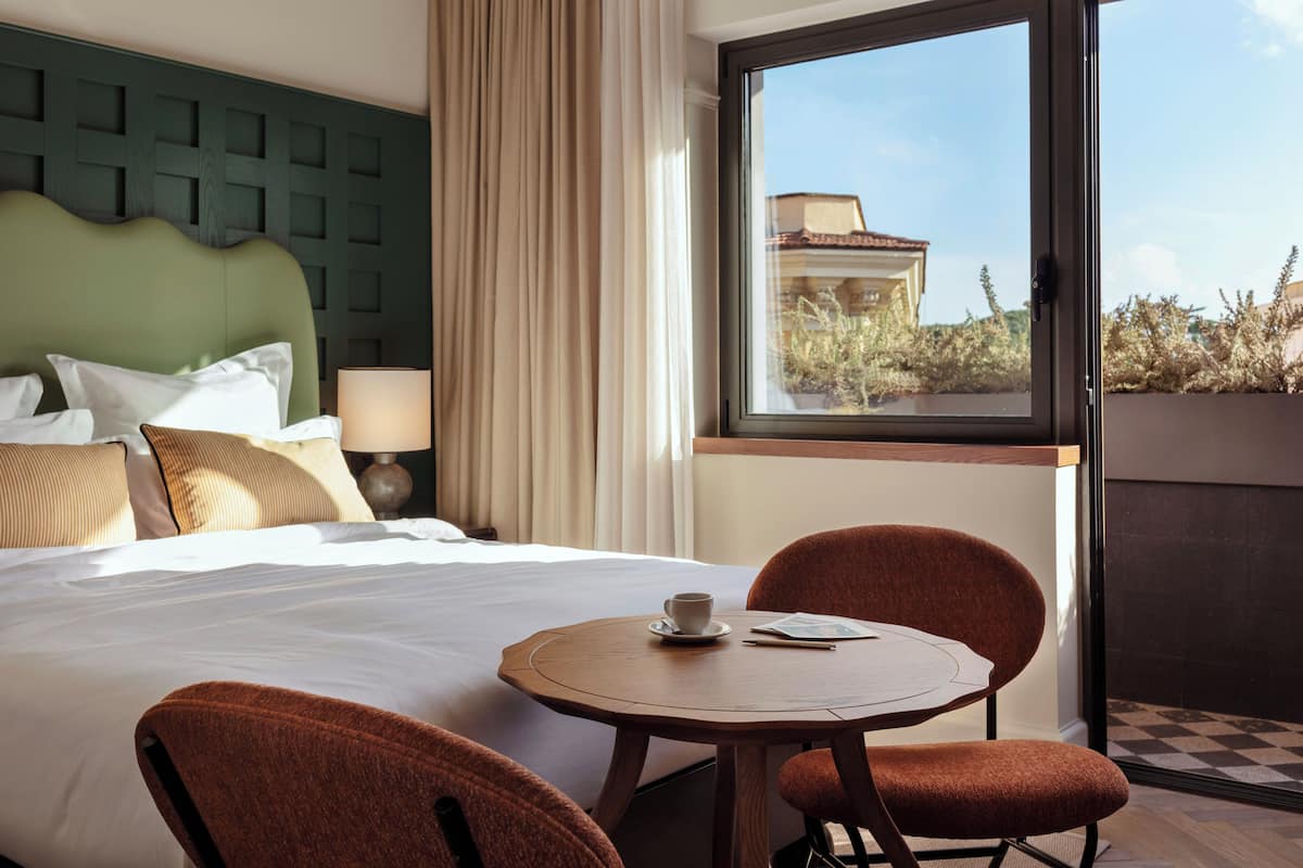 A hotel room with a large bed, a round table with two chairs, a view of the outside through a window, and a green headboard.
