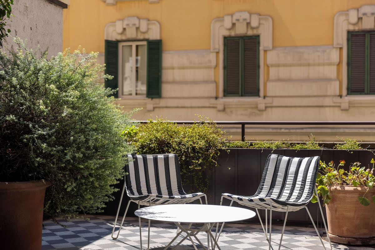 A checkered outdoor seating area with two striped chairs and a small table, surrounded by potted plants and a building with green shutters in the background.