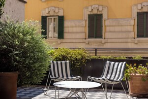A checkered outdoor seating area with two striped chairs and a small table, surrounded by potted plants and a building with green shutters in the background.