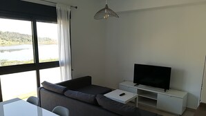 A modern living room with a sofa, a TV on a stand, a dining table, and a view of a lake through large windows.