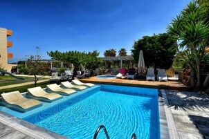 Una piscina con sillas de descanso y un tobogán, rodeada de palmeras y una zona de estar cubierta.