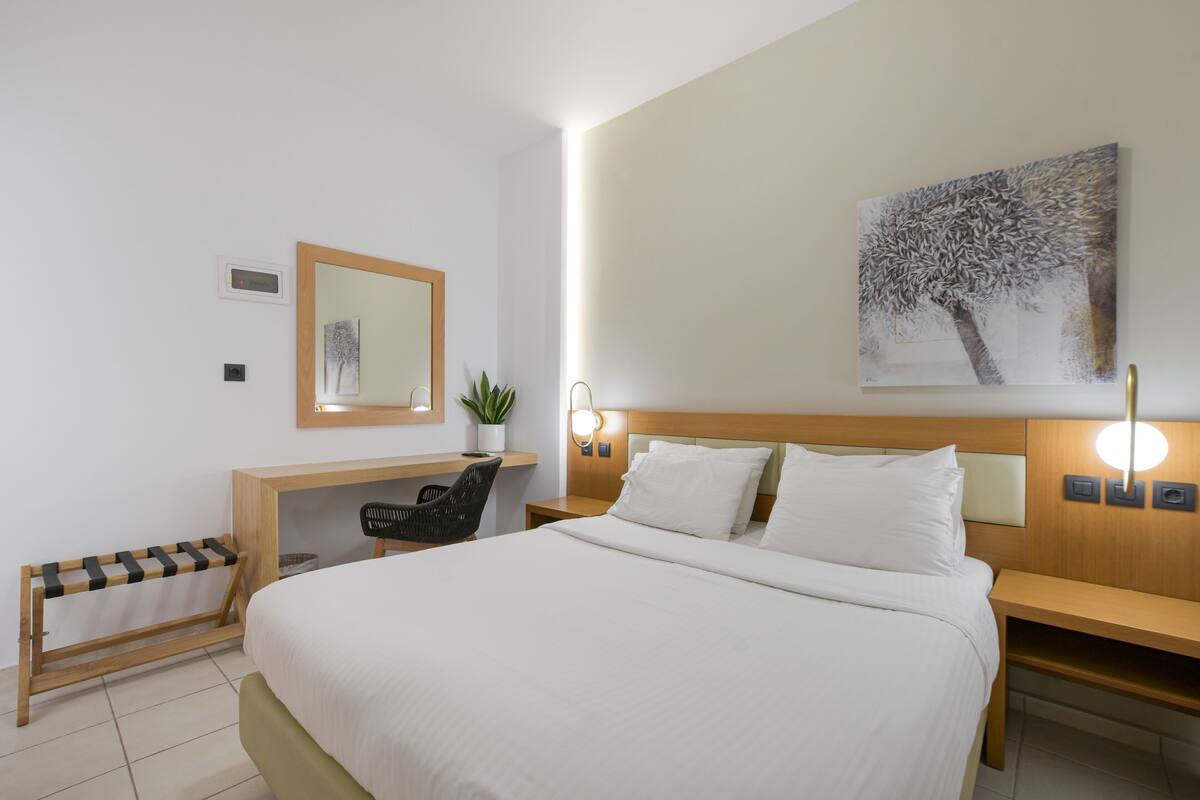 A hotel room with a large bed, a desk with a chair, a small plant, a wall-mounted mirror, and a painting.