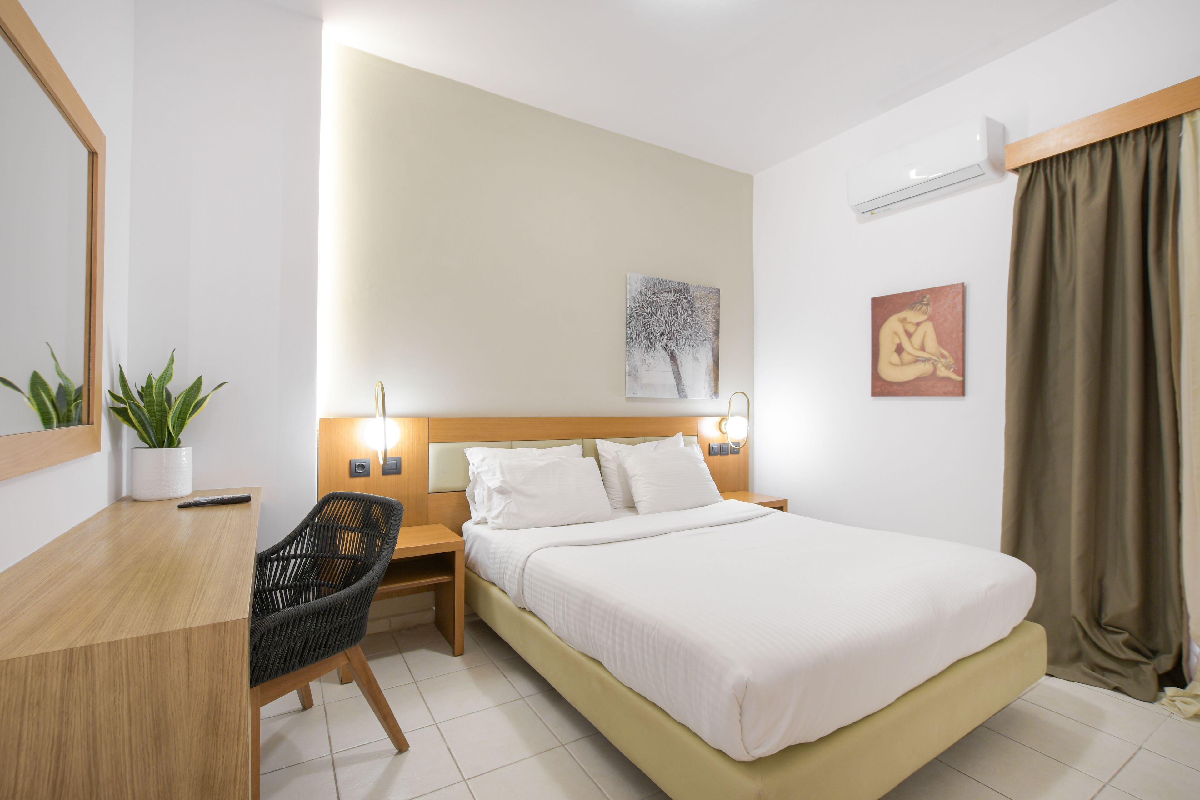 A hotel room with a bed, bedside table, chair, potted plant, artwork, and an air conditioning unit.