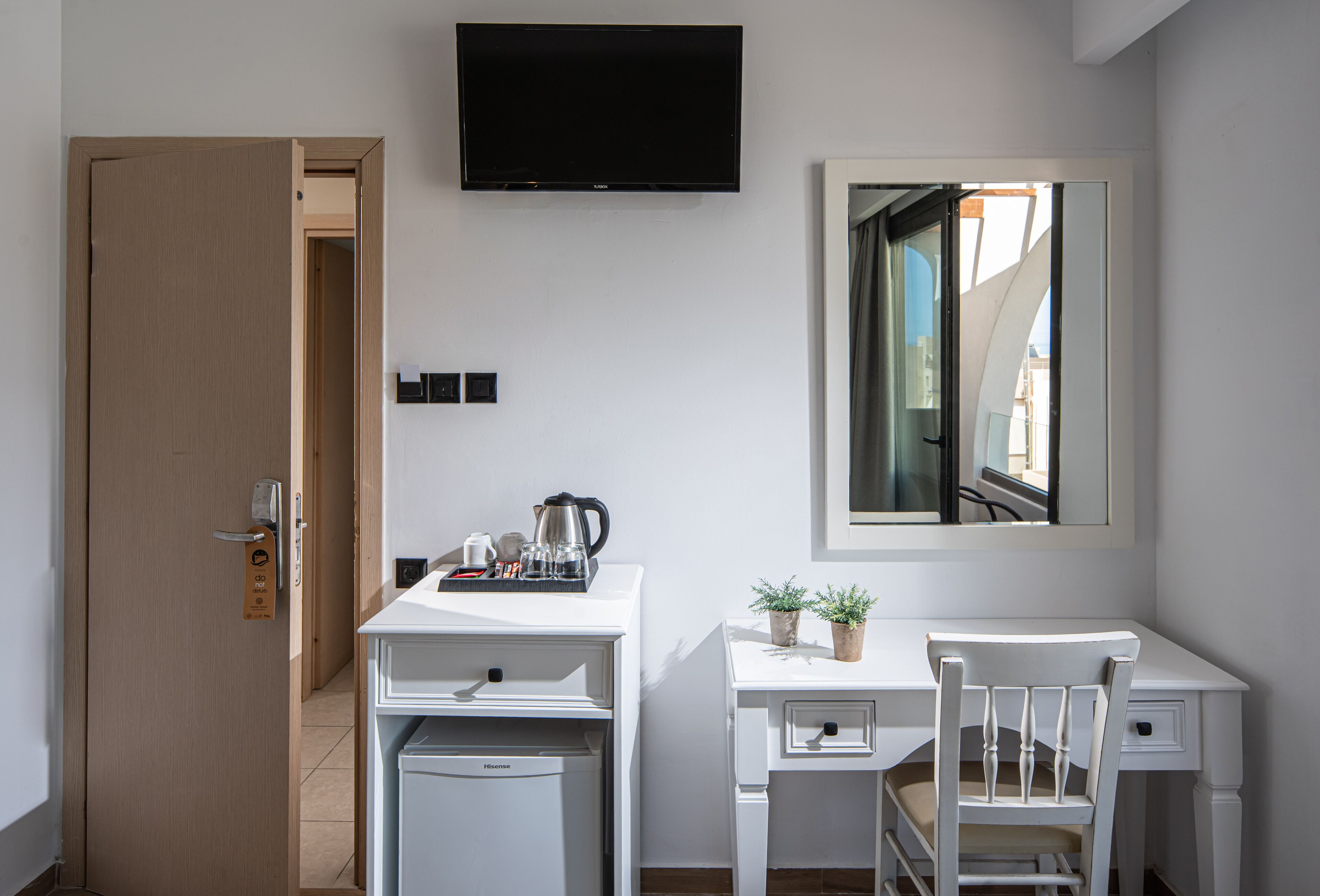 A compact hotel room with a kitchenette, a desk, a chair, a TV, and a mirror.