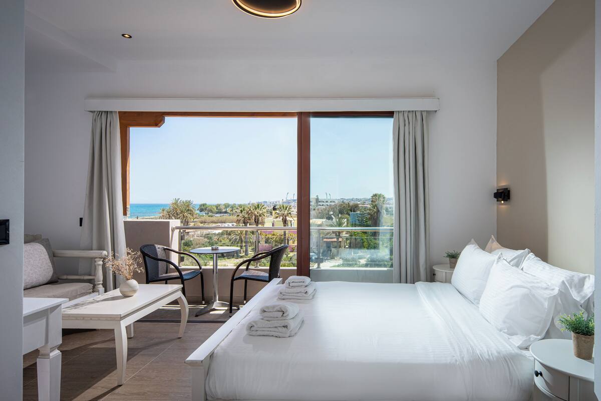 A hotel room with a large bed, a desk, a chair, and a balcony with a view of the sea.