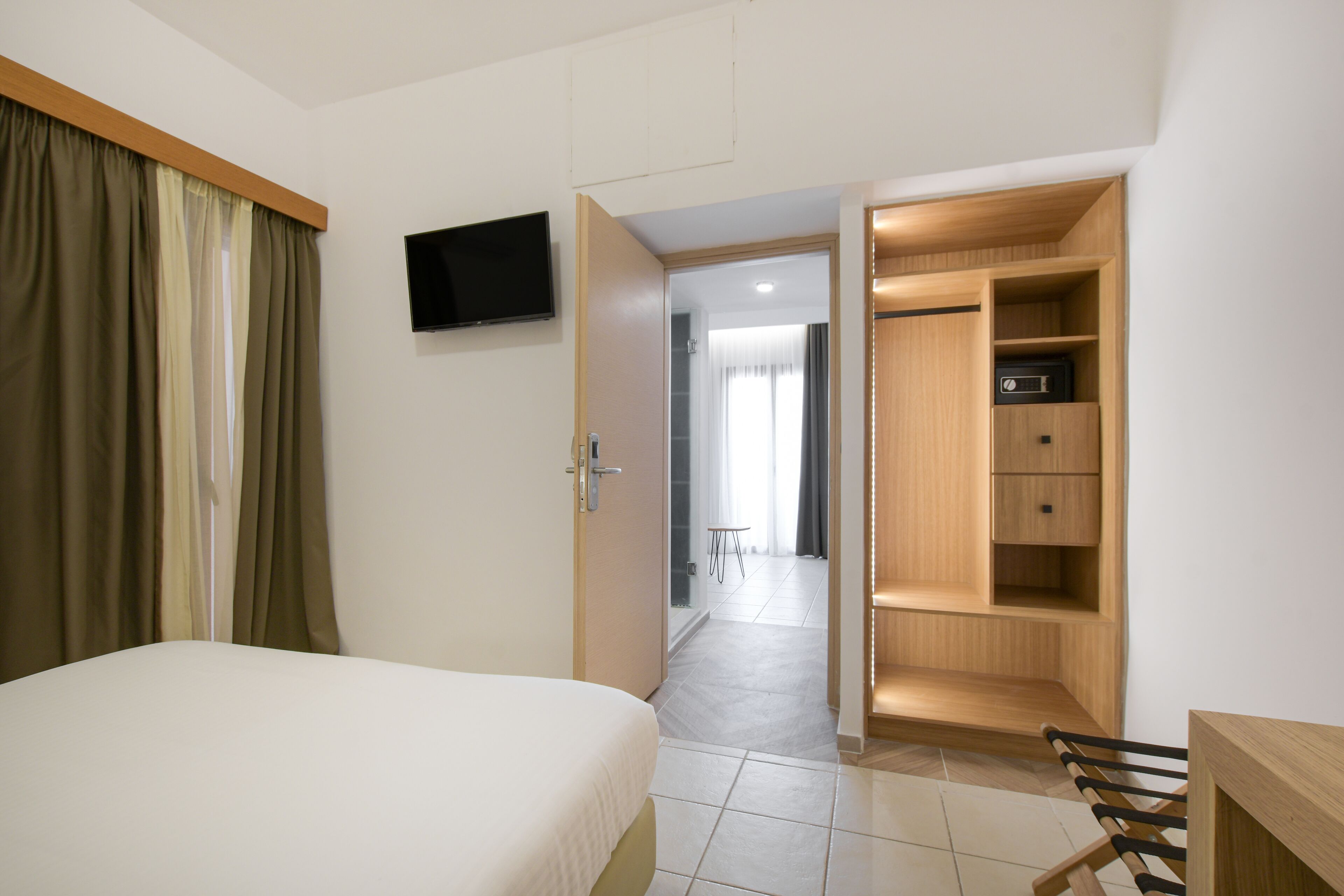 Habitación de hotel con cama, televisor montado en la pared, armario de madera y una mesita en el fondo.