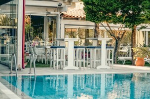 Un área de piscina al aire libre con taburetes y mesas blancas, rodeada de paredes de vidrio y plantas en macetas.