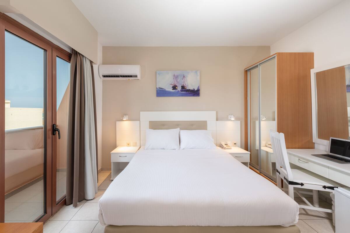 A hotel room with a large bed, a desk with a laptop, a painting on the wall, and an air conditioning unit.