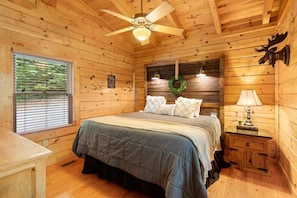 A wooden cabin bedroom with a bed, a nightstand, a lamp, a ceiling fan, and a window with blinds.
