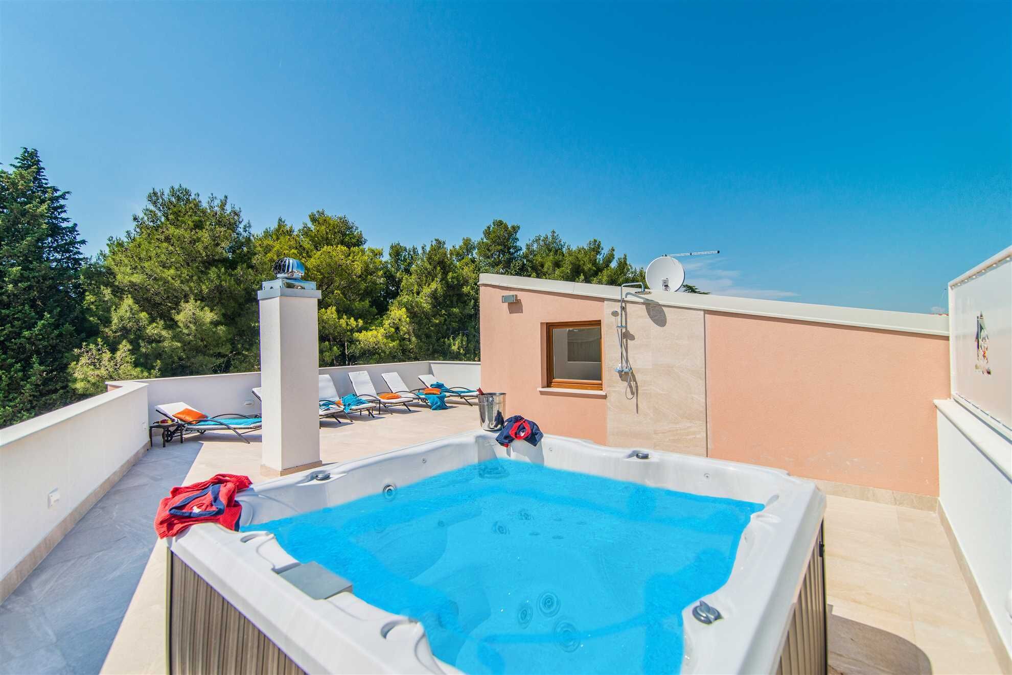 A rooftop terrace with a hot tub, lounge chairs, and a view of trees.