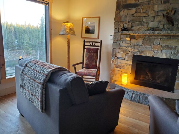 A cozy living room with a stone fireplace, a comfortable sofa, a chair, and a large window offering a view of a forest.