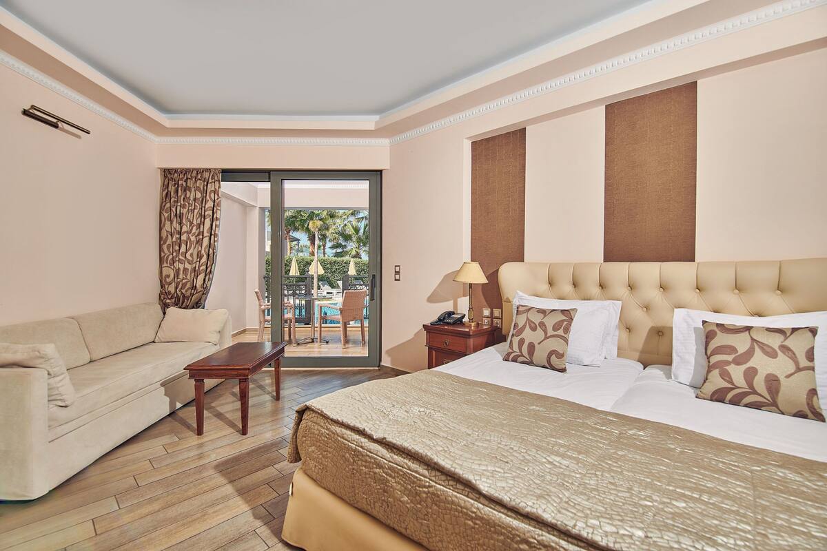 A hotel room with a bed, a sofa, a small table, and a view of an outdoor area with chairs and umbrellas.