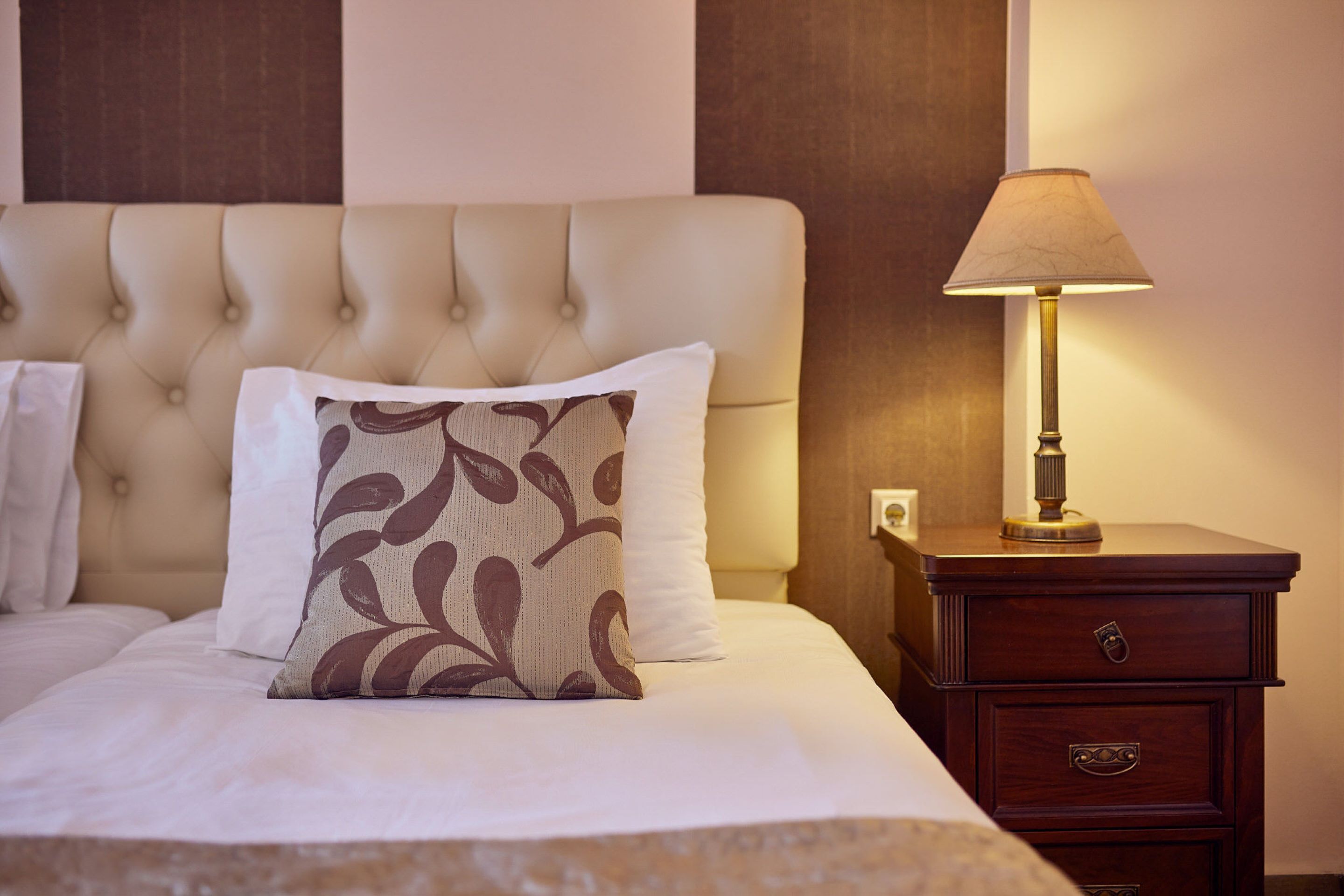 A neatly made bed with a tufted headboard, a patterned pillow, and a bedside lamp on a wooden nightstand.