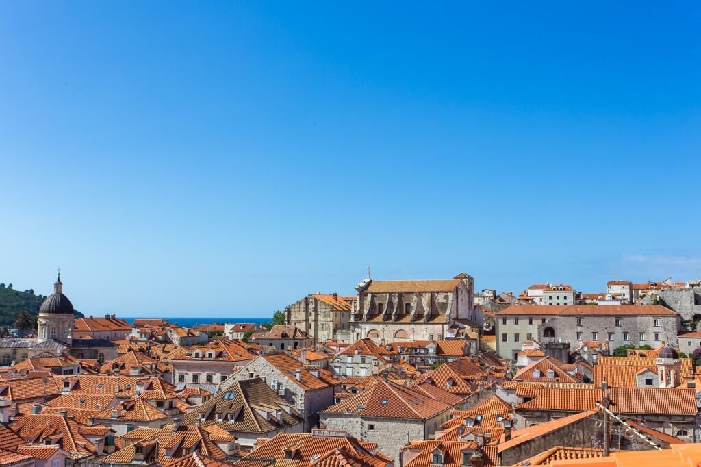 A historic cityscape with terracotta-roofed buildings, a prominent church with a dome, and a clear blue sky.