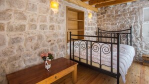 Un dormitorio con pared de piedra, vigas de madera en el techo, una cama con estructura metálica, una mesita de noche y un ventanal pequeño.
