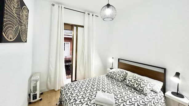 A bedroom with a bed, a bedside table with a lamp, a radiator, and a door with curtains.