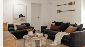 A modern living room with two dark sofas, a round coffee table, a framed painting, and two wooden paddles on the wall.