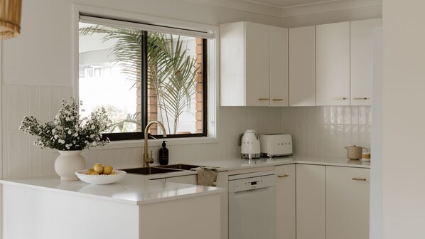 A modern kitchen with white cabinetry, a built-in dishwasher, and a window with a view of palm trees.